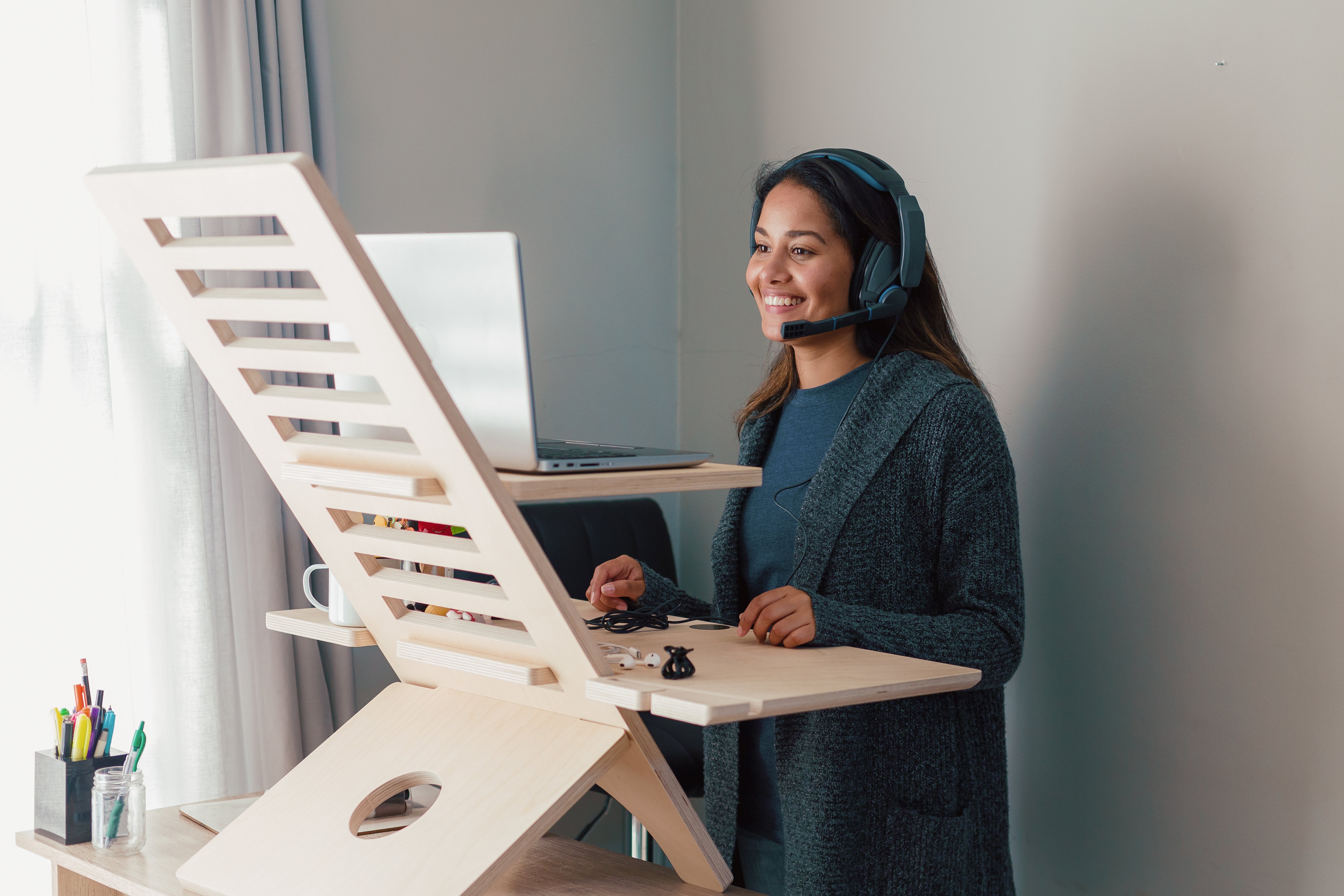 Young woman with long black hair and wearing black headphones working at a wooden standing desk facing a laptop in her home office